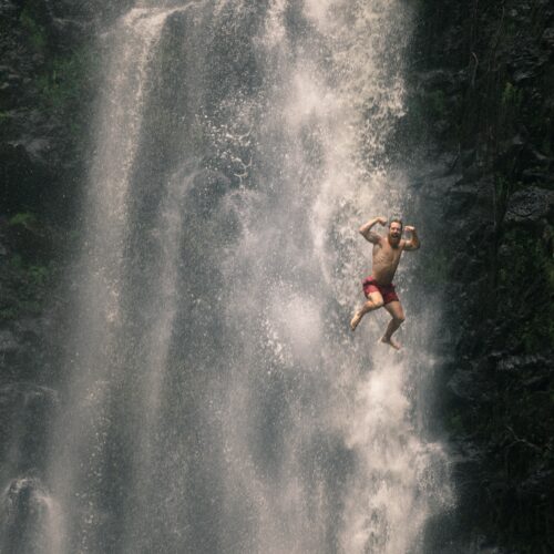 A man in swimming trunks has jumped from the top of a waterfall and he is pictured in mid-flight in front of the waterfall