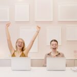 Two girls sitting at a desk with open laptops and they both look very happy. One girl has her arms raised above her head in jubilation