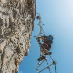 A man holds onto a rope ladder on the side of a cliff face with blue sky above