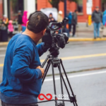 A man stands behind a video camera filming someone on the other side of the road