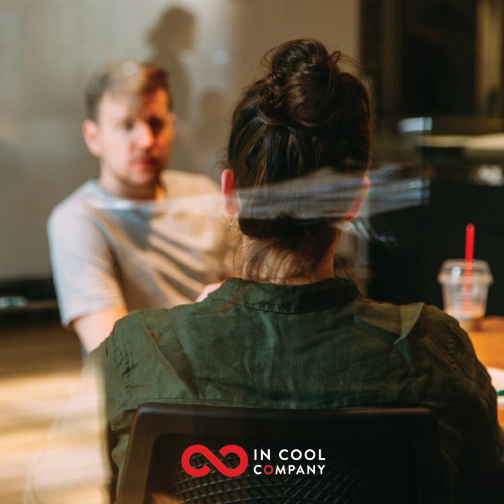 A man dressed casually sits talking to a woman across a table