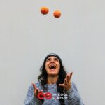 A man with long hair, wearing a beany hat is juggling two apples and smiling widely