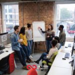 A small group of people stand around a flip chart in an office for a sales meeting
