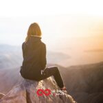 A female sitting on a mountain looking out at a sunset over a lake