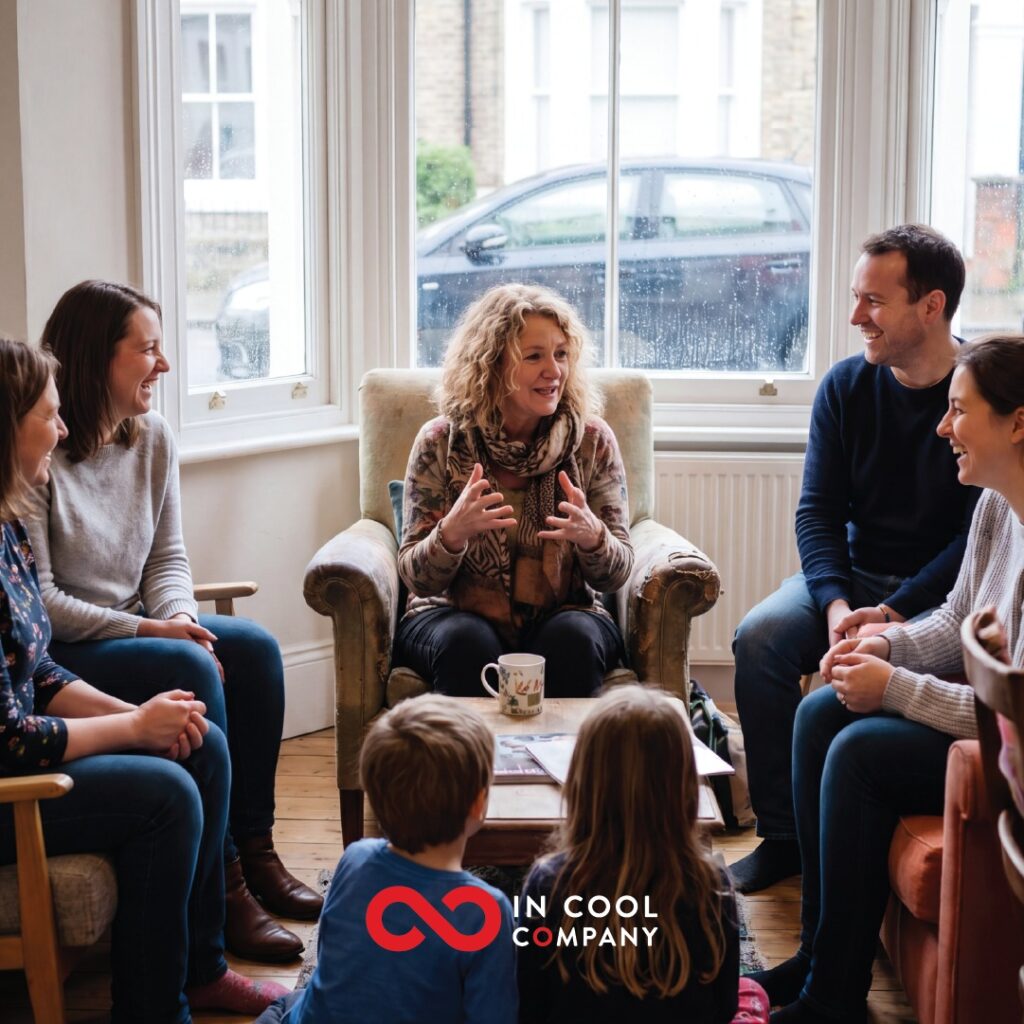 A lady sits in a comfy chair with other on chairs around her and 2 children on the floor. Everyone is looking at the lady as she talks
