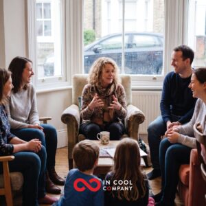 A lady sits in a comfy chair with other on chairs around her and 2 children on the floor. Everyone is looking at the lady as she talks
