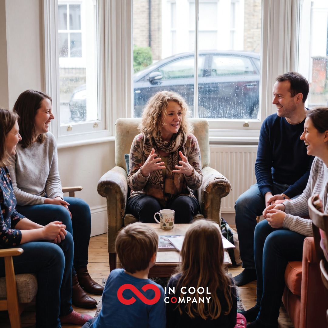 A lady sits in a comfy chair with other on chairs around her and 2 children on the floor. Everyone is looking at the lady as she talks