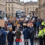 A small group of people stand in a street with placards listening to a man talk on a small stage