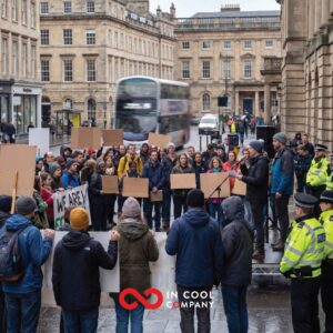 A small group of people stand in a street with placards listening to a man talk on a small stage