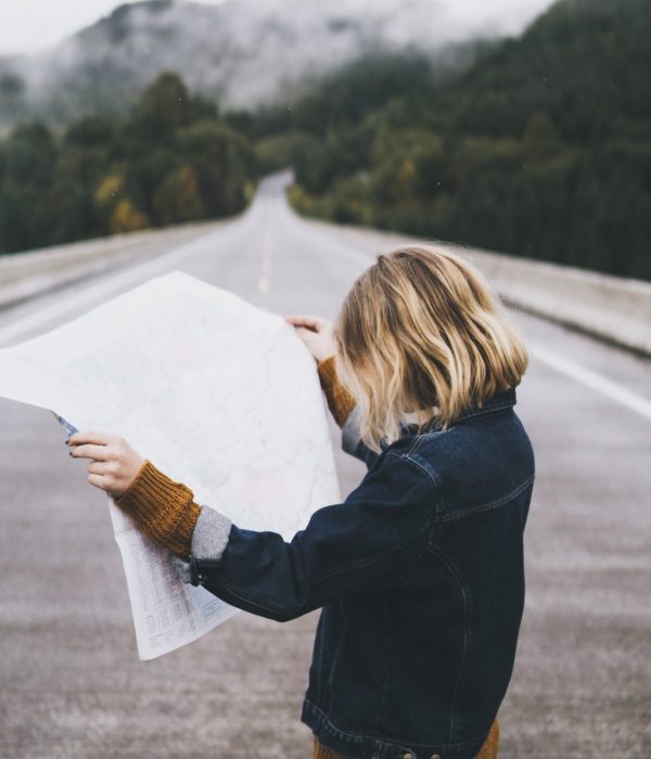 Woman in denim jacket standing in the middle of an empty road with forests in the background. The woman is holding a large paper map and studying it